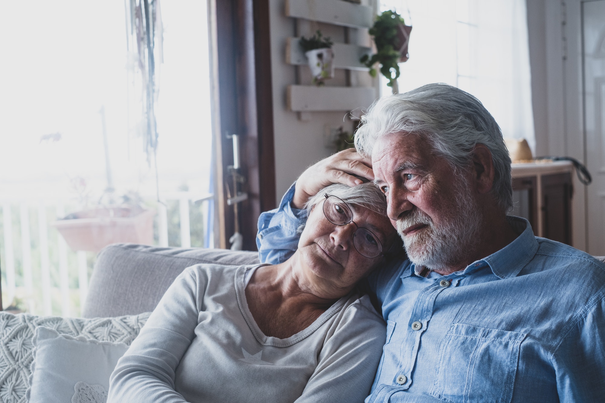 Couple of two sad and depressed old people sitting on the sofa at home looking outside the window.