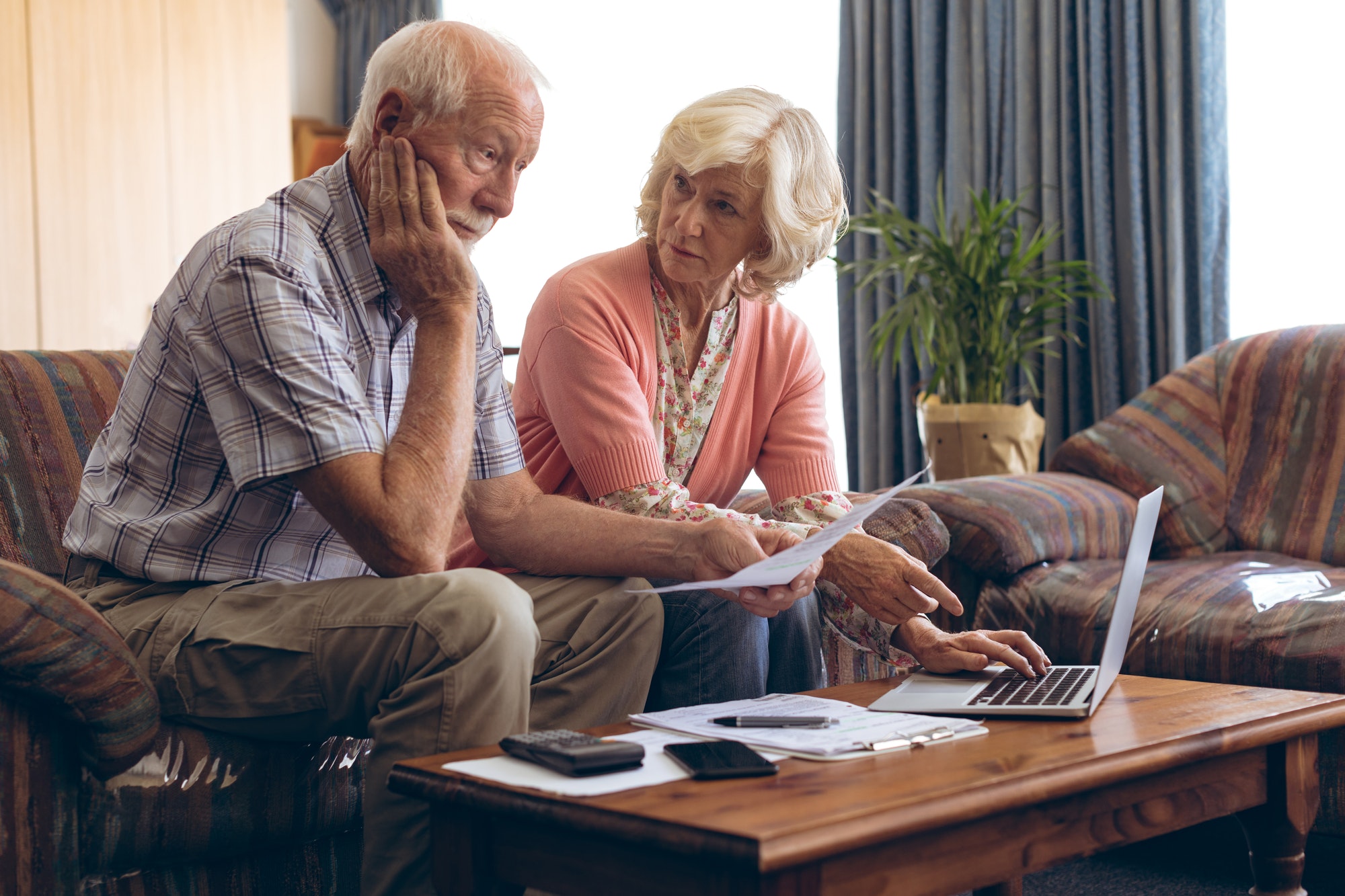 Sad senior couple discussing bill while sitting on vintage sofa at retirement home